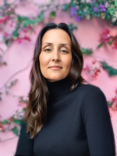 The image shows a woman with long dark brown hair wearing a black turtleneck sweater. She has a calm expression on her face, and the background features soft pink hues with blurred flowers and greenery.